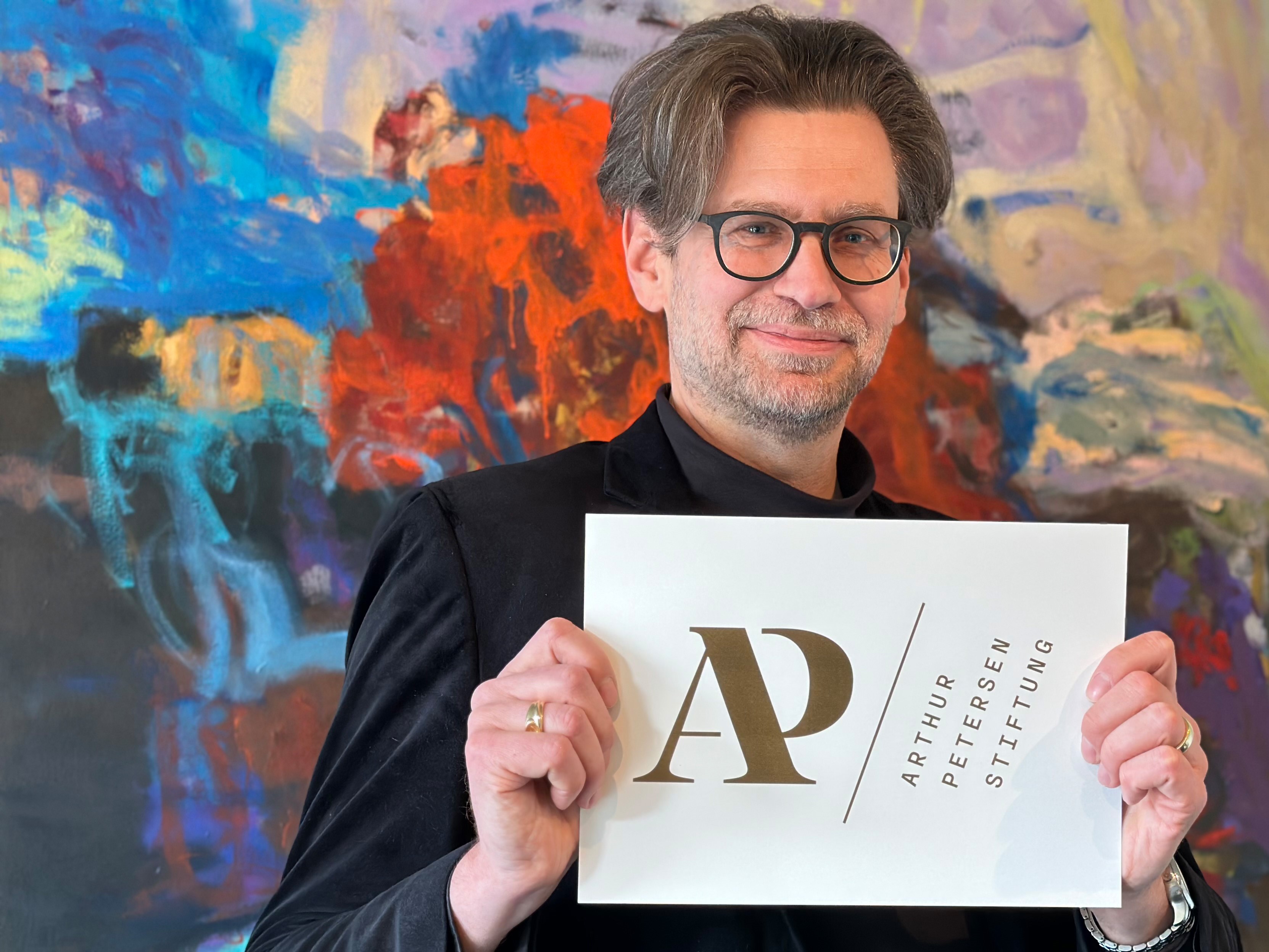 Das Foto zeigt Arne Zerbst, Präsident der Kunsthochschule, der ein Logo in der Hand hält. Darauf zu lesen ist "AP" - Arthur-Petersen-Stiftung.