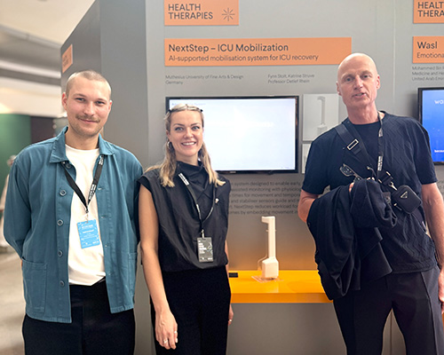 Drei Personen stehen in einer Ausstellung und pr&auml;sentieren ein Designprojekt