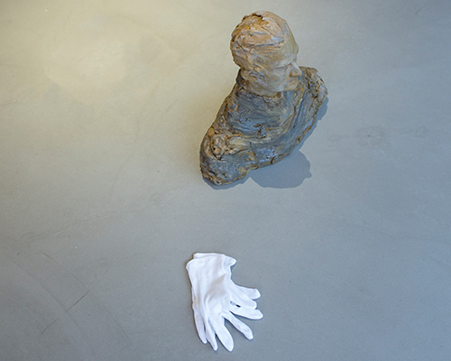 Das Bild zeigt eine Büste, die auf einem Boden steht. Daneben liegen weiße Handschuhe.