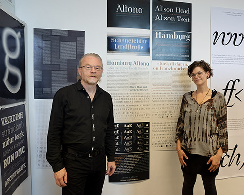 Das Bild zeigt zwei Personen, die vor Straßenschildern und Schrift-Drucken stehen
